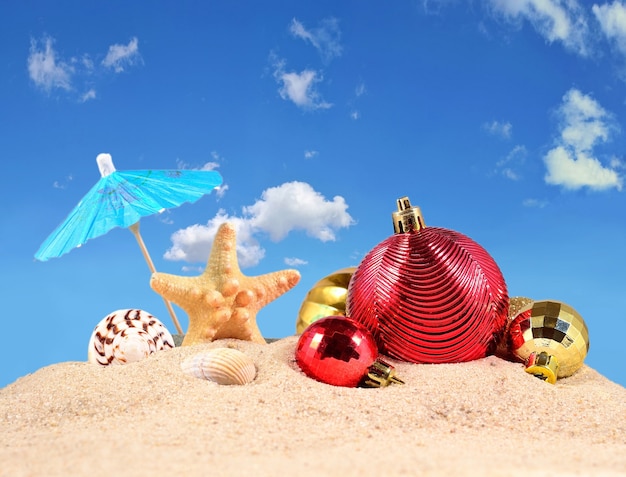 Adornos navideños, conchas y estrellas de mar en la arena de una playa contra el cielo azul