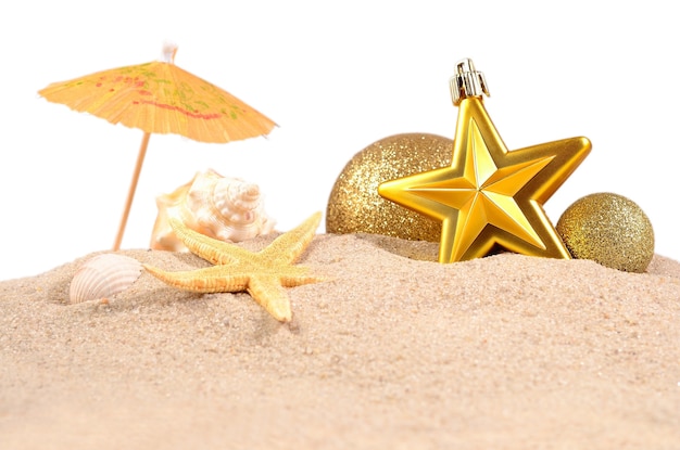 Adornos navideños con conchas y estrellas de mar en la arena de una playa sobre un fondo blanco.