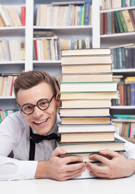 Adoro estudar. Jovem alegre de camisa e gravata borboleta, sentado à mesa da biblioteca e abraçando uma pilha de livros