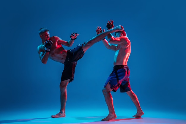 Adrenalin. MMA. Zwei professionelle Kämpfer, die auf blauem Studiohintergrund in Neon stanzen oder boxen. Fit muskulöse kaukasische Athleten oder Boxer kämpfen. Sport, Wettbewerb und menschliche Emotionen, Anzeige.