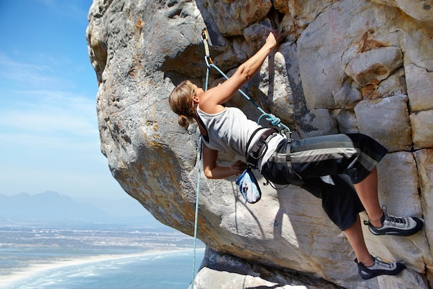 La adrenalina la está empujando hacia la cima Una mujer joven escalando una pared de roca mientras está enmarcada contra un cielo azul