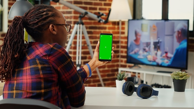 Adulto afro-americano segurando verticalmente smartphone com tela verde, sentado na mesa de casa. Trabalhando com modelo de simulação de espaço de cópia em branco e exibição de chave croma isolada. Tiro de tripé.