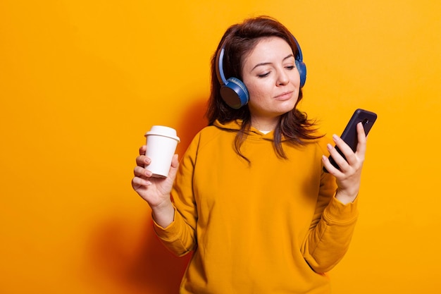 Adulto caucasiano, segurando o telefone celular e a xícara de café enquanto usa fones de ouvido para ouvir a música. Mulher alegre curtindo música e áudio no fone de ouvido enquanto usa o smartphone e bebe