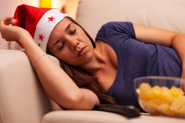 Adulto com chapéu de Papai Noel dormindo no sofá após assistir filme de entretenimento de dezembro