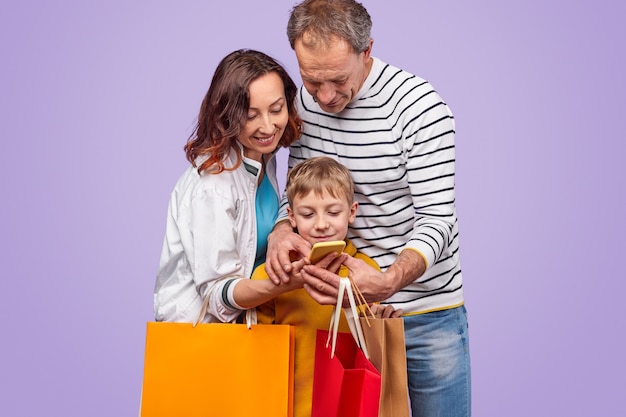 Adulto e filho alegres com sacos de papel navegando no smartphone juntos em pé contra um fundo roxo e fazendo compras