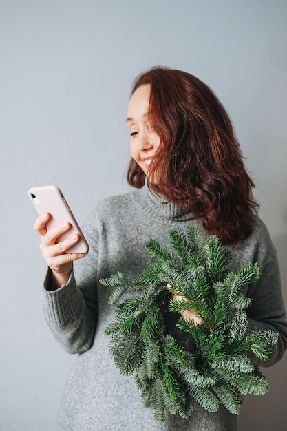 Adulto hermosa mujer sonriente cuarenta años con cabello rizado morena en vestido de punto gris cálido con corona de Navidad de abeto diy en la mano usando teléfono móvil sobre fondo gris