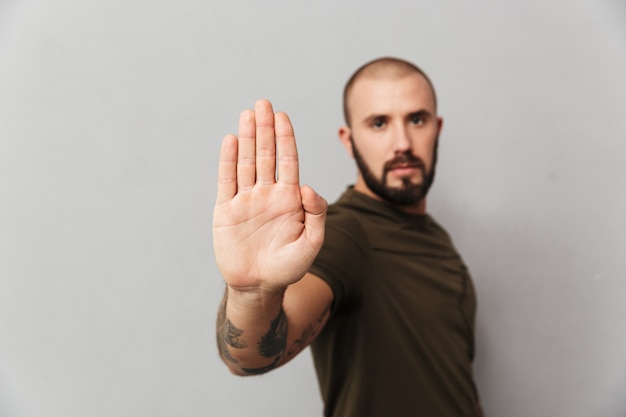 Adulto homem bonito com tatuagens nas mãos, posando e mostrando o gesto de parada com a mão, isolada sobre parede cinza