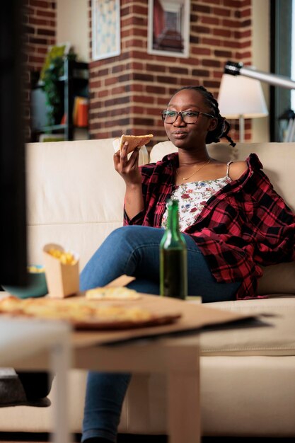 Un adulto joven alegre comiendo un trozo de pizza y viendo una película, relajándose en el sofá con entrega de comida para llevar. Disfrutando de la comida rápida del paquete de comida para llevar para ver películas en la televisión.