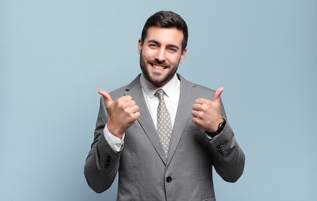 Foto adulto joven apuesto hombre de negocios sonriendo con alegría y luciendo feliz, sintiéndose despreocupado y positivo con ambos pulgares hacia arriba