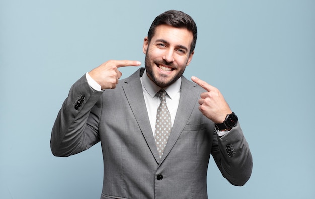 Foto adulto joven apuesto hombre de negocios sonriendo con confianza apuntando a su propia sonrisa amplia, actitud positiva, relajada y satisfecha