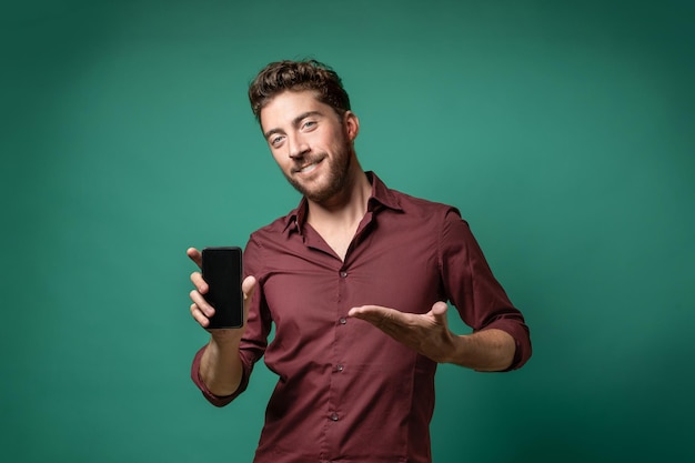 Adulto joven barbudo en camisa de color rojo oscuro que muestra la pantalla del teléfono inteligente aislado contra un fondo verde