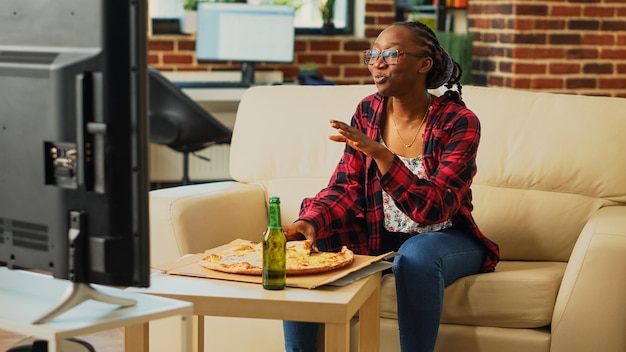 Adulto joven comiendo una rebanada de pizza en la sala de estar, disfrutando de una película de acción en el canal de televisión. Grl relajado sintiéndose feliz bebiendo una botella de alcohol y cenando comida para llevar, programa de televisión.