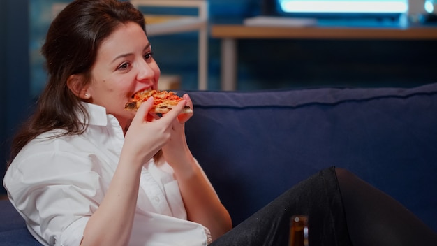 Adulto joven disfrutando de una rebanada de pizza y cerveza sentado en el sofá