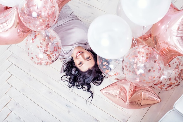 Adulto joven hermosa mujer en vestido de fiesta de moda con globos de color rosa acostado sobre un piso de madera