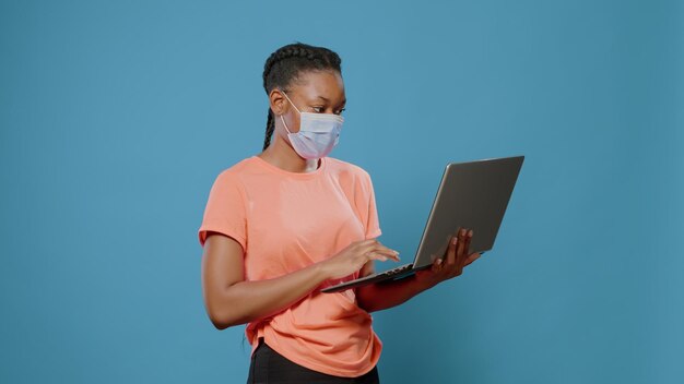 Adulto joven con mascarilla con portátil moderno en estudio. Mujer que trabaja con tecnología en una computadora inalámbrica durante la pandemia de coronavirus. Persona de moda mirando la pantalla del dispositivo.