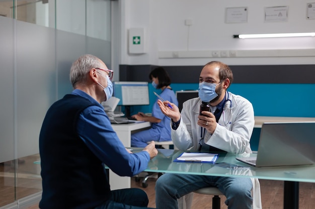 Adulto jubilado que recibe un frasco de píldoras del médico en la consulta durante la pandemia de covid 19. Médico con mascarilla dando tratamiento con receta en frasco al paciente en la visita de control.