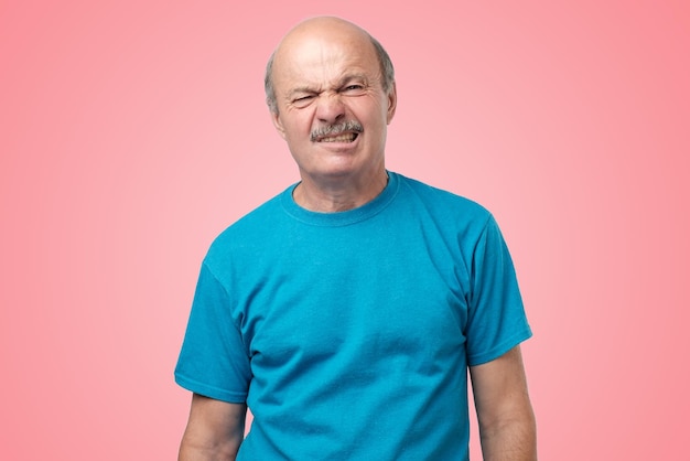 Adulto maduro em camiseta azul com dúvida com nojo no rosto em pé no fundo rosa
