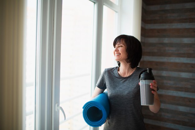 Adulto slim mulher tem treino em casa. Fique de pé após o treino com um pouco de proteína e tapete de ioga nas mãos. Preocupe-se com a boa forma e o bem-estar do corpo.