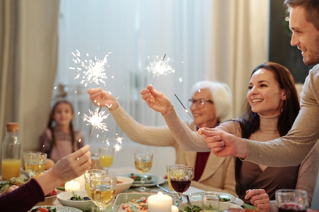 Adultos jóvenes y maduros alegres sosteniendo brillantes luces de bengala sobre la mesa festiva servida durante la cena de Navidad