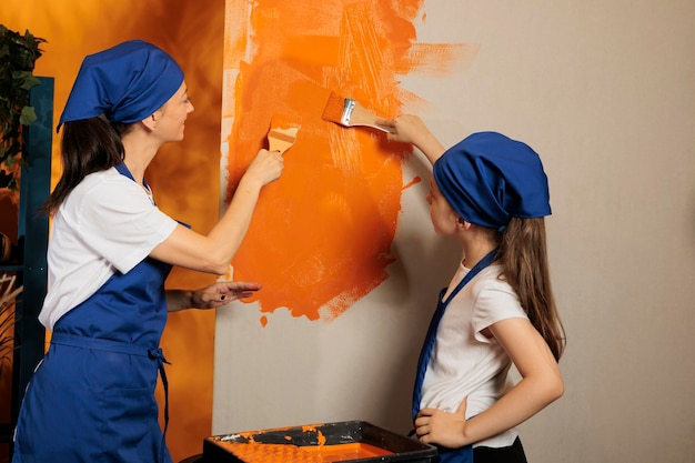 Adultos y niños pintando paredes naranjas con pintura acrílica y pincel, utilizando herramientas y equipos renovadores para la decoración interior del hogar. Familia haciendo trabajo en equipo redecorando apartamento.