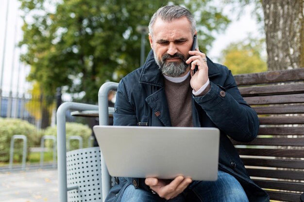Advogado adulto maduro com barba trabalha usando um laptop e fala ao telefone na rua