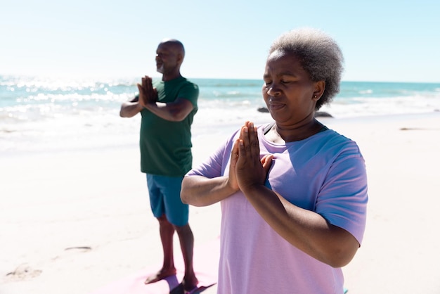 Ältere afroamerikanische Männer und Frauen meditieren im Gebet am Strand vor klarem Himmel. Kopierraum, Sommer, unverändert, Liebe, zusammen, Yoga, Ruhestand, Natur, Urlaub und aktiver Lebensstil.