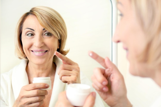 Foto Ältere frau, die kosmetische creme auf gesicht nahe spiegel aufträgt