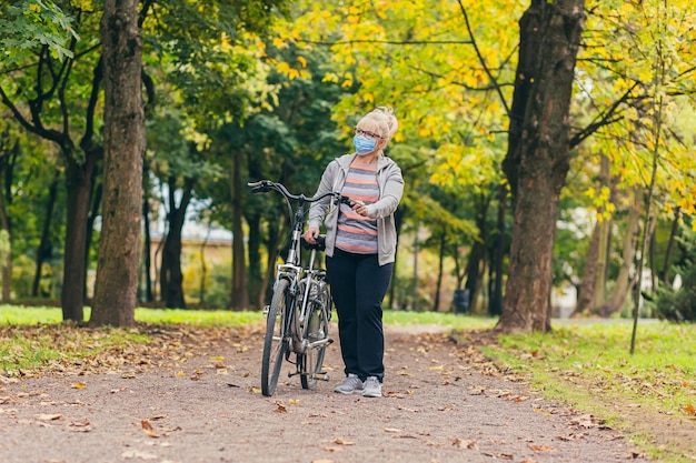 ältere Frau geht im Park mit einem Fahrrad in einer medizinischen Schutzmaske