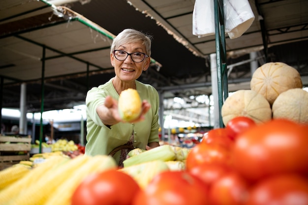 ältere Frau kauft Mais und Obst auf dem Marktplatz und hält eine Tasche voller gesunder Lebensmittel