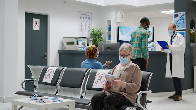 Ältere frau mit gesichtsmasken, die darauf warten, an einem kontrollbesuch im empfangsbereich des krankenhauses teilzunehmen, virenschutz. patienten, die während einer coronavirus-pandemie im wartezimmer einer medizinischen klinik sitzen.