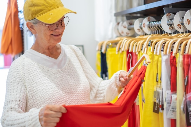 Ältere Frau mit Hut und Brille im modernen Laden, die bunte neue Kleidung auswählt