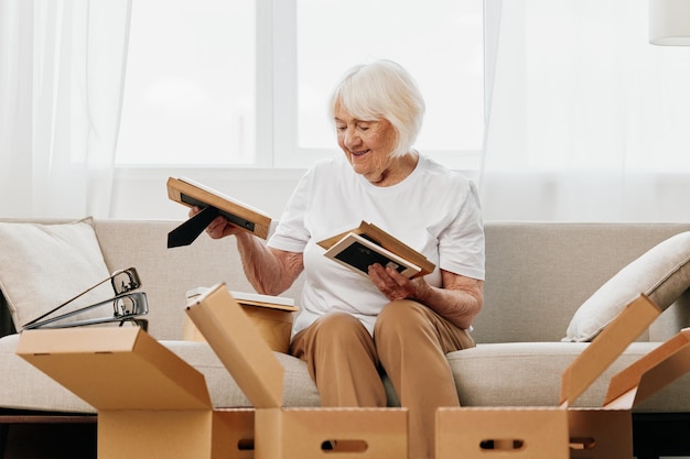 Ältere Frau sitzt zu Hause auf einem Sofa mit Kisten, sammelt Dinge mit Erinnerungen, Alben mit Fotos und Fotorahmen, zieht an einen neuen Ort und putzt Dinge und ein glückliches Lächeln. Lebensstil im Ruhestand