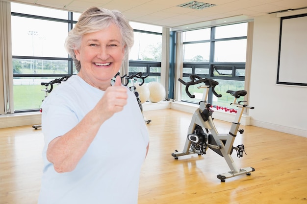 Foto Ältere frau zeigt daumen hoch gegen sich drehende heimtrainer im fitnessraum