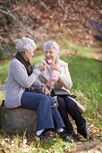 Foto Ältere frauen glücklich und snack im park mit süßigkeiten zahnseide essen und zusammen zu entspannen auf dem ruhestand im freien senior freunde lächeln und junk food auf urlaub auf dem land und bindung in der natur