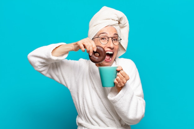 Ältere hübsche frau nach der dusche und im bademantel und beim frühstück mit kaffee und donut