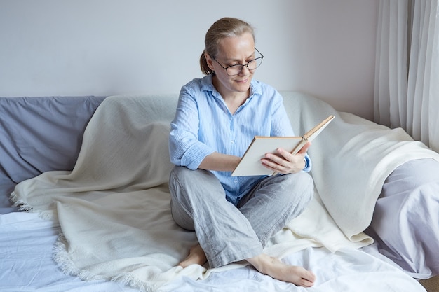 Ältere lächelnde frau mit brille sitzt mit ihren beinen auf dem sofa, liest ein buch.