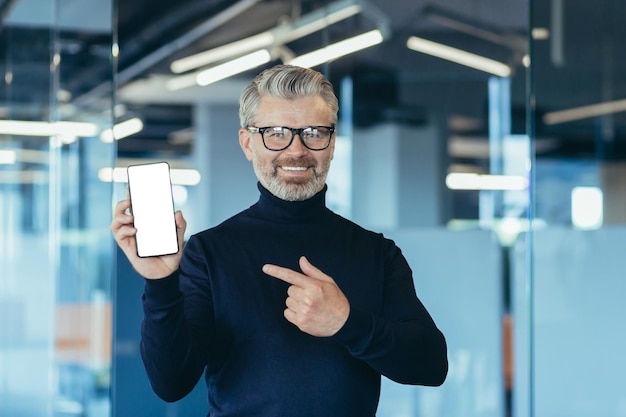 Älterer erfolgreicher grauhaariger geschäftsmann im büro, der telefonbildschirm zur kamera mit weißem bildschirm zeigt