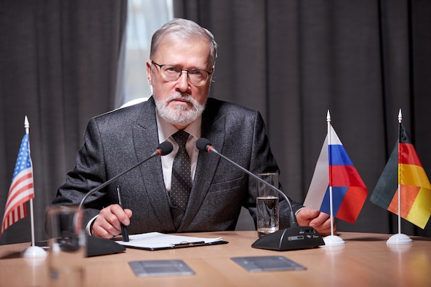 Foto Älterer geschäftsmann, der mit mikrofon im modernen sitzungssaal während des treffens mit geschäftspartnern sitzt, gutaussehender mann in brille, der zuversichtlich spricht, formellen anzug tragend