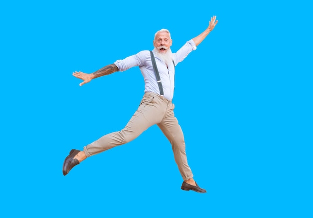 Älterer Hipster-Mann mit Tätowierung, der Spaß beim Springen auf blauem Hintergrund hat, glücklicher Senioren-Lebensstil