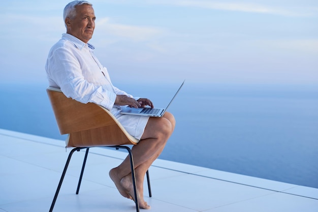 älterer Mann, der an einem Laptop arbeitet und den Sonnenuntergang mit Meerblick auf dem Balkon eines modernen Hauses betrachtet