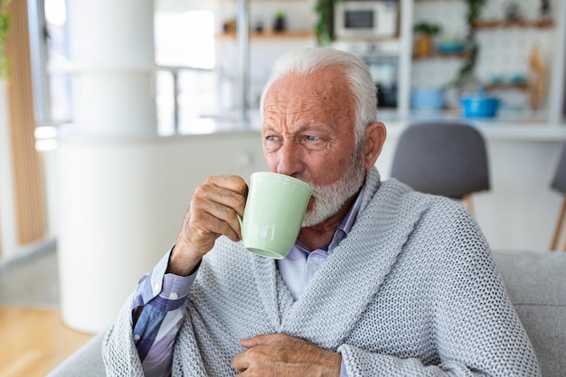 Älterer Mann, der an Grippe leidet und Tee trinkt, während er in eine Decke gehüllt auf dem Sofa sitzt