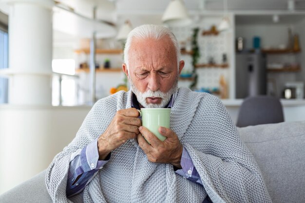 Älterer Mann, der an Grippe leidet und Tee trinkt, während er zu Hause in eine Decke gehüllt auf dem Sofa sitzt Kranker Mann mit einer Erkältung, der auf dem Sofa liegt und eine Tasse heißen Tee hält
