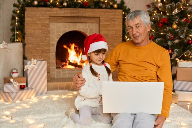 Älterer mann im gelben hemd, das auf boden sitzt und laptop auf knien hält, seine enkelin umarmt und ameisenkamera sieht, niedliches kind, das notebook-bildschirm betrachtet, kleidet weihnachtsmütze.