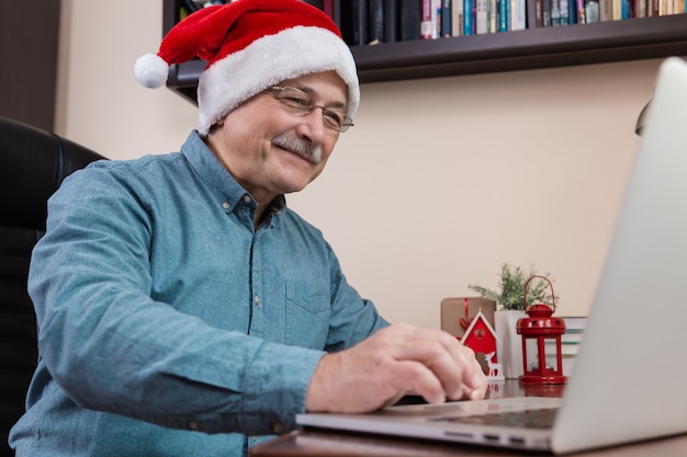 Älterer mann im weihnachtsmannhut spricht unter verwendung des laptop-geräts für videoanruffreunde und -kinder. das zimmer ist festlich eingerichtet.