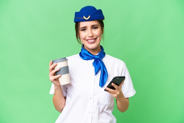 Aeromoça de avião sobre fundo croma isolado segurando café para levar e um celular