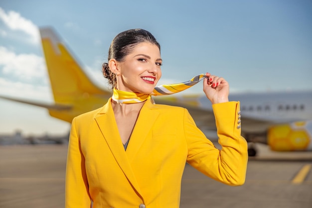 Aeromoça feminina alegre com cachecol de aeromoça no pescoço, olhando para a câmera e sorrindo em pé ao ar livre no aeródromo