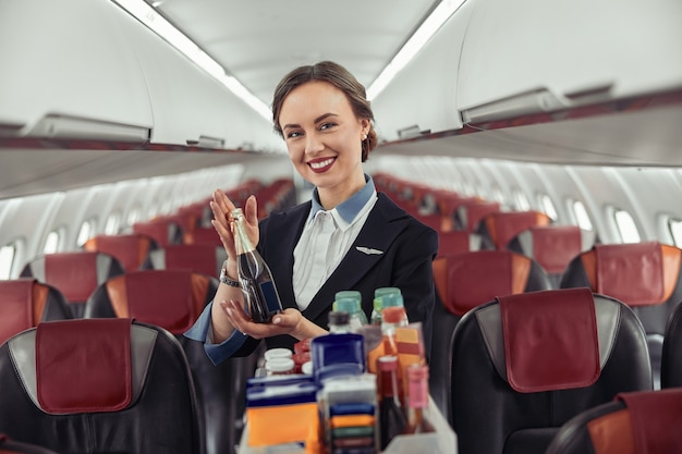 Aeromoça mostra garrafa com álcool na cabine de passageiros do avião a jato. Interior moderno do avião. Mulher europeia sorridente usa uniforme e olhando para a câmera. Aviação comercial civil. Conceito de viagem aérea