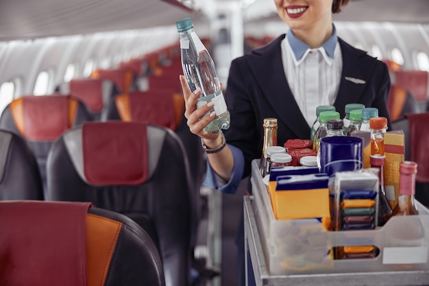 Aeromoça segurando a garrafa de água na cabine de passageiros do avião a jato. Interior moderno do avião. Imagem recortada de uniforme de mulher. Aviação comercial civil. Conceito de viagem aérea