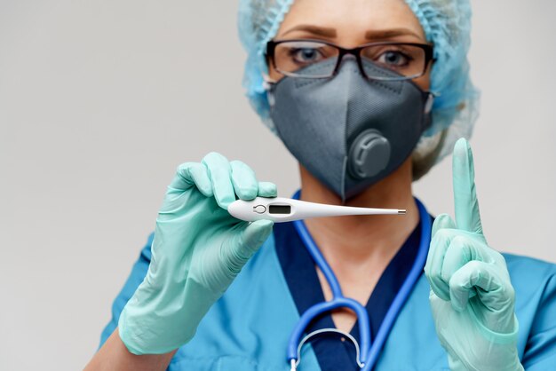 Ärztin mit Stethoskop tragen Schutzmaske und Latexhandschuhe über hellgrauem Hintergrund, der Thermometer hält