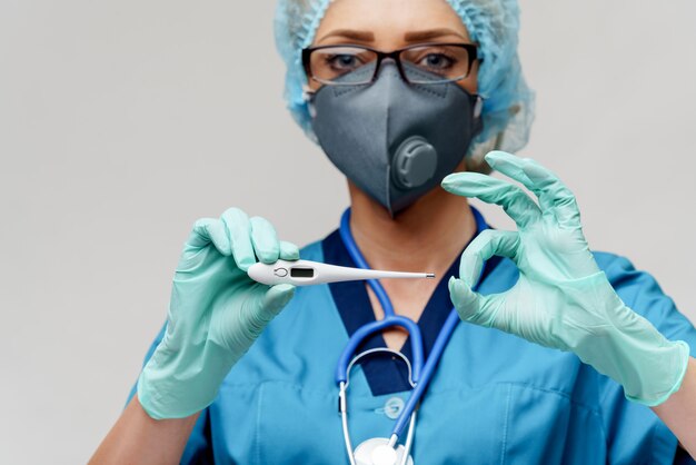 Ärztin mit Stethoskop tragen Schutzmaske und Latexhandschuhe über hellgrauem Hintergrund, der Thermometer hält